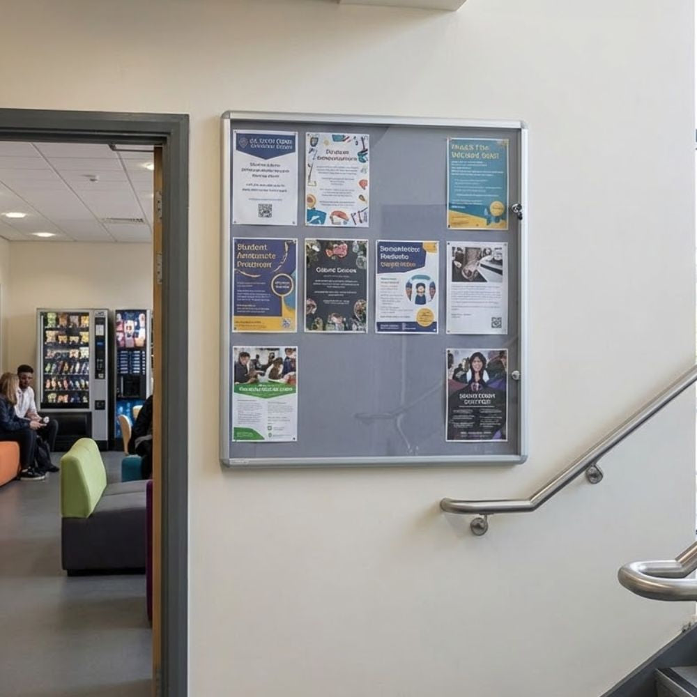 1200 x 1200mm fire resistant tamperproof noticeboard with grey felt surface and aluminium frame, installed on a stairwell wall in an education building displaying posters behind a secure lockable cover.