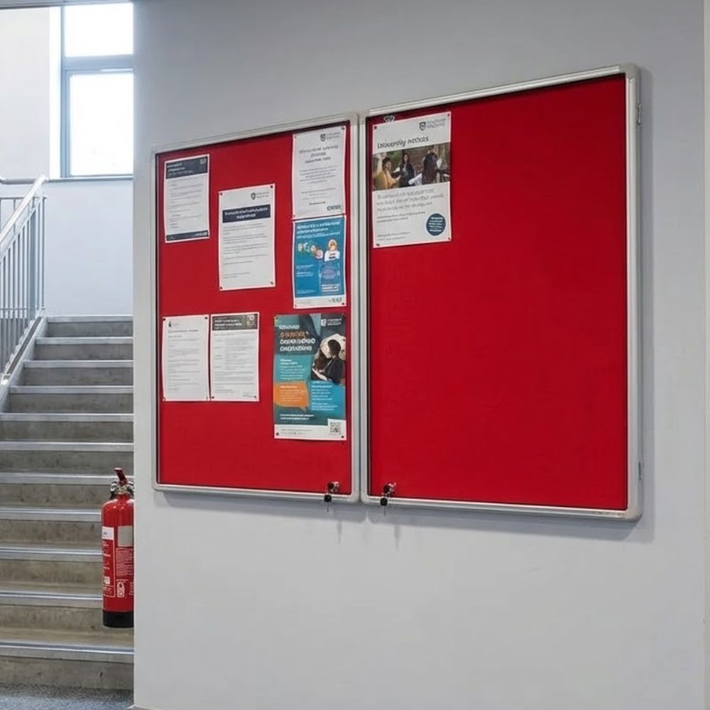 2400 x 1200mm fire resistant tamperproof noticeboard with red felt backing and aluminium frame, wall mounted in a stairwell corridor displaying official notices behind lockable glazed doors.