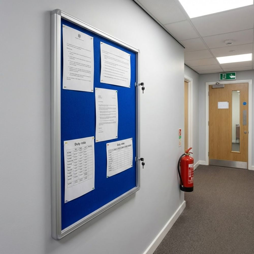 600 x 900mm fire resistant tamperproof noticeboard with blue felt backing and aluminium frame, wall mounted in a corridor displaying pinned documents behind a lockable glazed door.