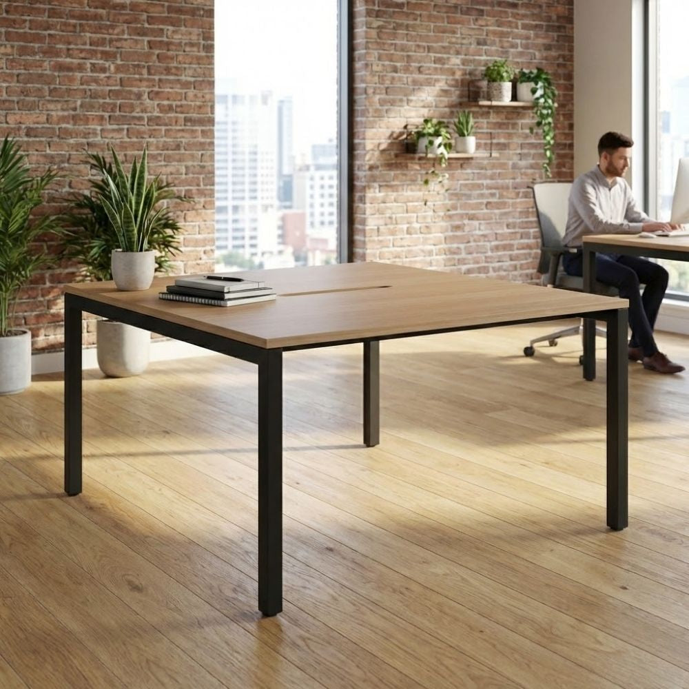 Bench-style back-to-back desk with black frame and central cable access slot, styled with notebooks and plants, placed in a modern office with exposed brick and wooden flooring.
