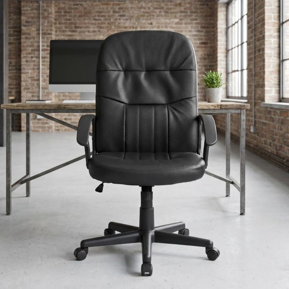 Black leather manager's office chair featuring fixed arms and smooth-rolling castors, positioned in front of a rustic wood and metal desk in an industrial-style workspace with exposed brick walls.