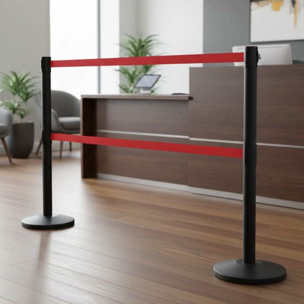 Black steel retractable barrier post with twin 3.4m red belts for high security queue management in a modern reception area with wooden flooring and desk in the background.