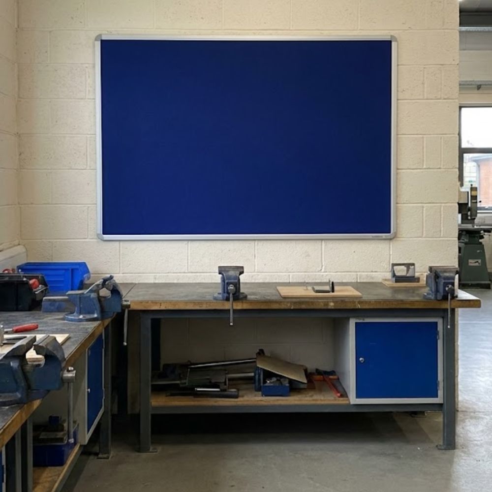 Blue aluminium framed noticeboard 1800 x 1200mm fixed to a brick wall above a workshop bench with tools and vices in an industrial setting.
