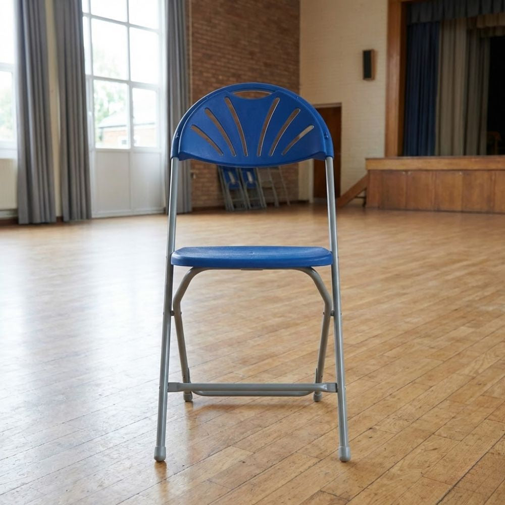 Blue plastic folding assembly chair with grey metal frame, positioned on a wooden floor in a school hall with stage and curtains in the background.