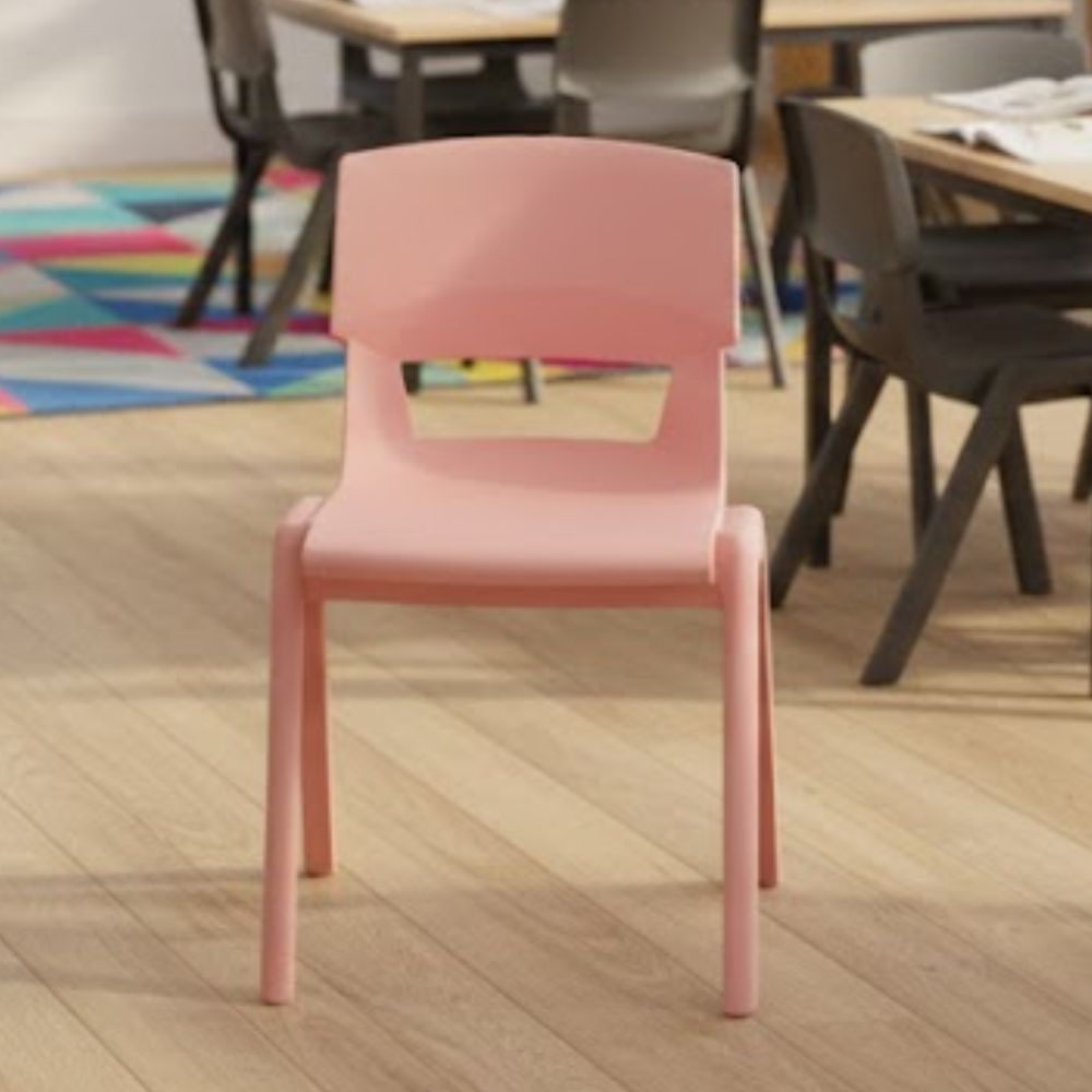 A blush pink school chair with an ergonomic backrest, set within a classroom of mixed-colour chairs and group tables on light timber flooring.