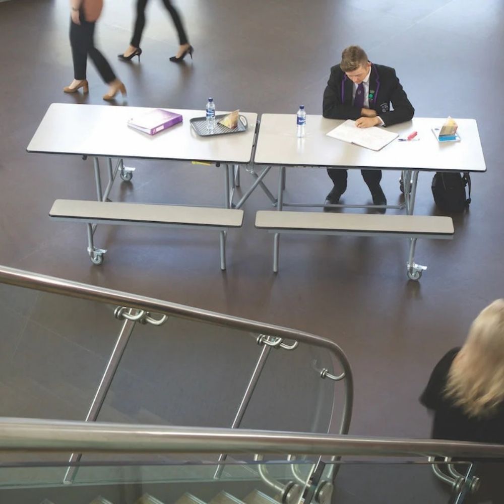 Student using a white canteen bench table in a school dining hall setting with folding frame and seating benches.