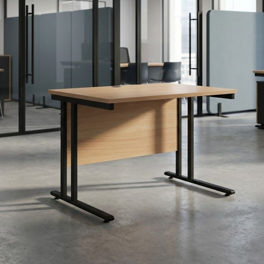 Compact rectangular office desk with a wood-finish top, black cantilever frame, and modesty panel, placed in a modern workspace with concrete flooring and glass partitions.
