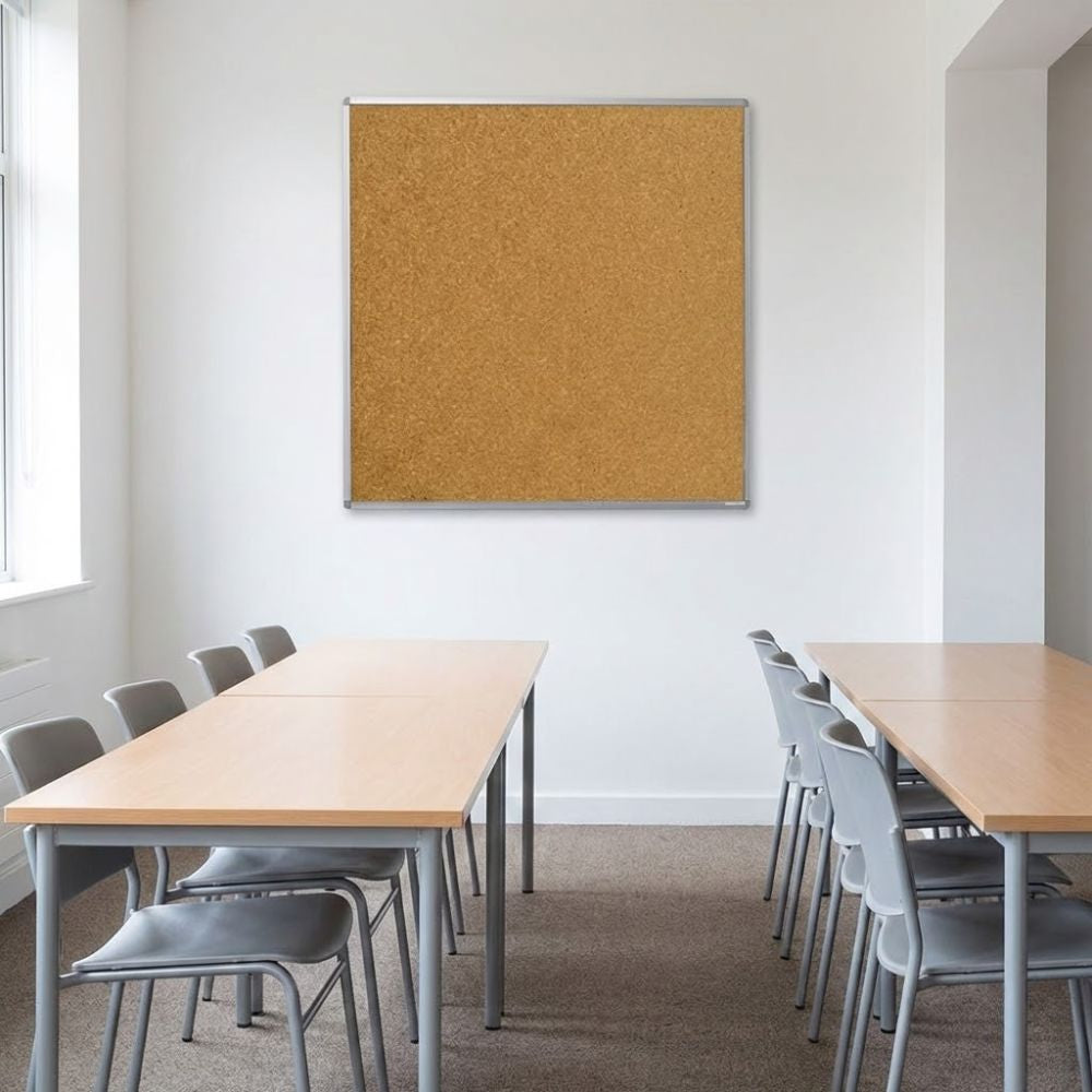 Cork aluminium framed noticeboard 1200 x 1200mm fixed to a white wall in a meeting room with rectangular tables and grey chairs arranged below.