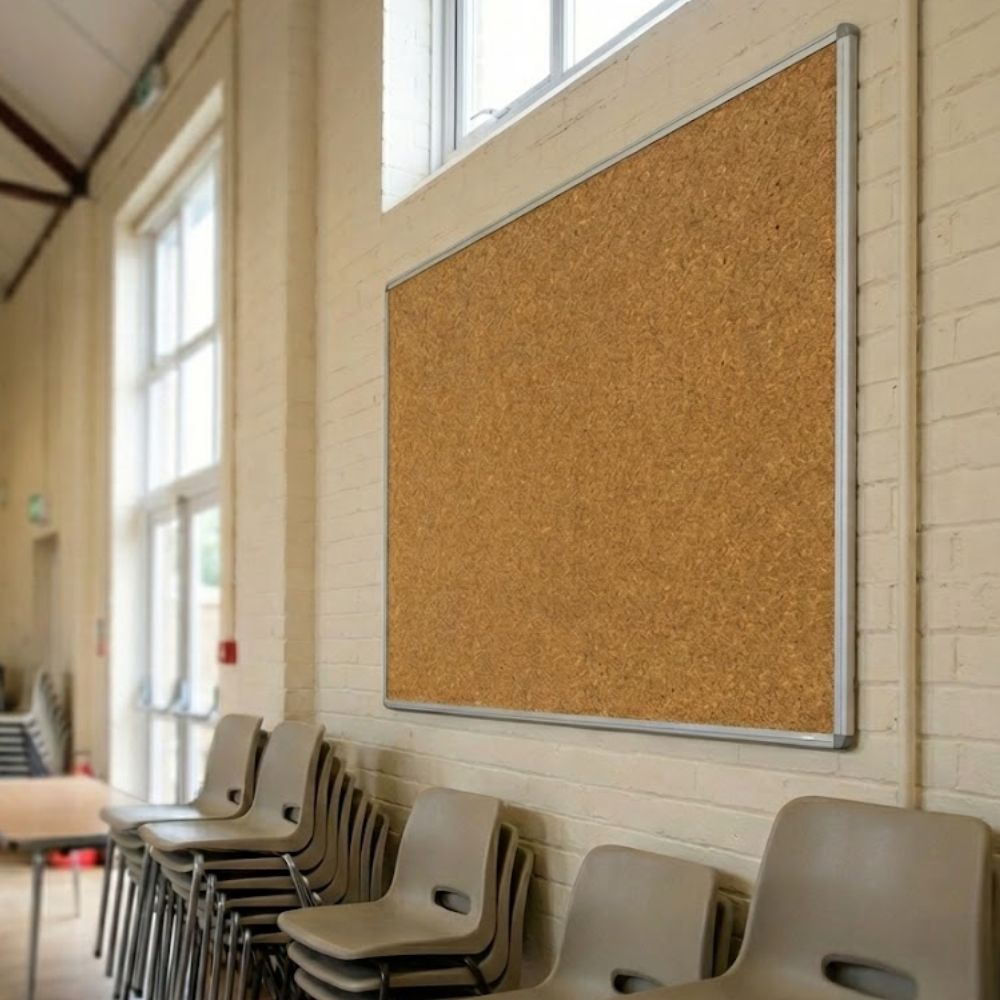 Cork aluminium framed noticeboard measuring 1500 x 1200mm fixed to a brick wall in a hall with stacked chairs positioned beneath it.