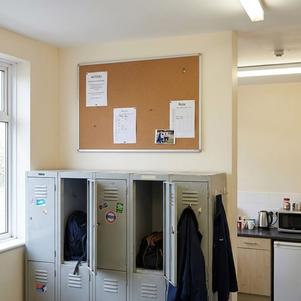 Cork aluminium framed noticeboard fixed to a wall above metal lockers in a staff room with kitchen facilities and pinned notices.