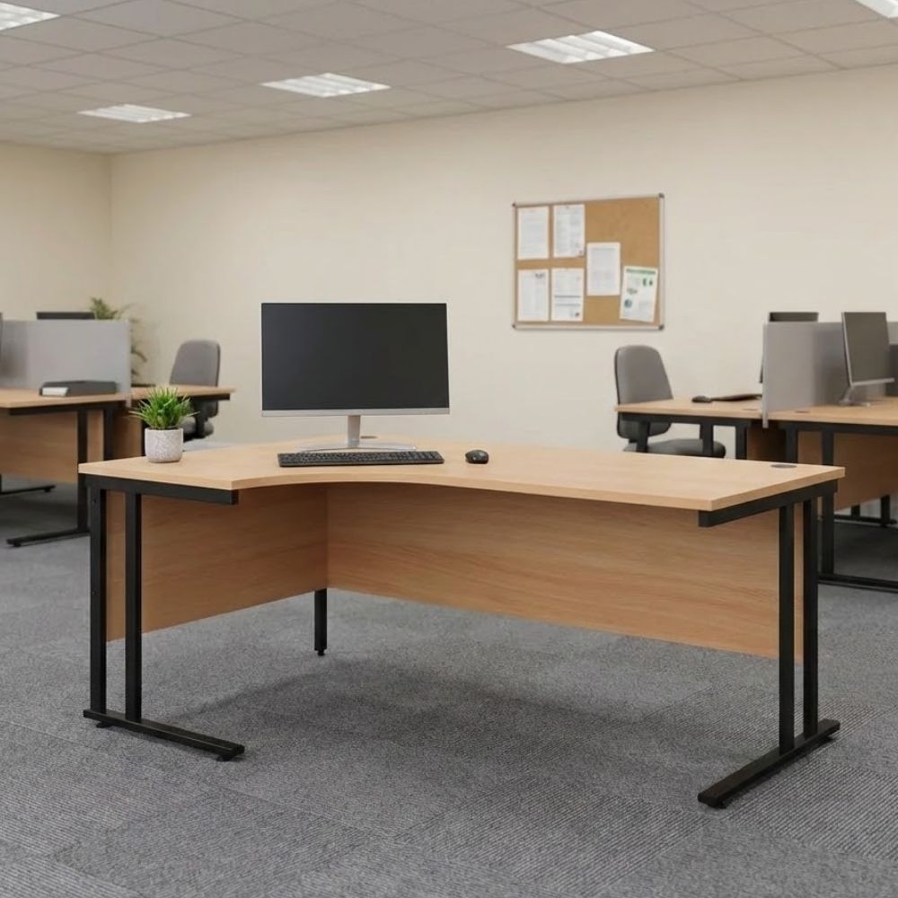 Spacious left-hand return corner desk featuring a wood-finish surface, modesty panel, black metal frame, and desktop monitor, located in a carpeted open-plan office.