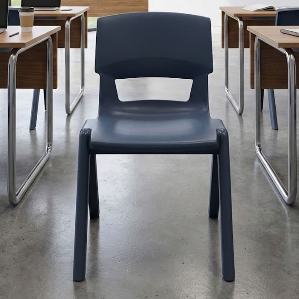 A dark grey plastic classroom chair in a minimalist study environment, placed between modern wood-and-metal desks on polished concrete flooring.