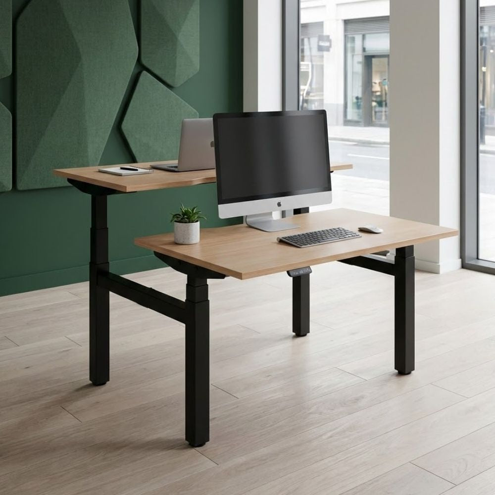Back-to-back electric sit-stand desks with black frames and natural wood tops, each configured at different heights, positioned in a bright office with full-height windows and green acoustic panels.