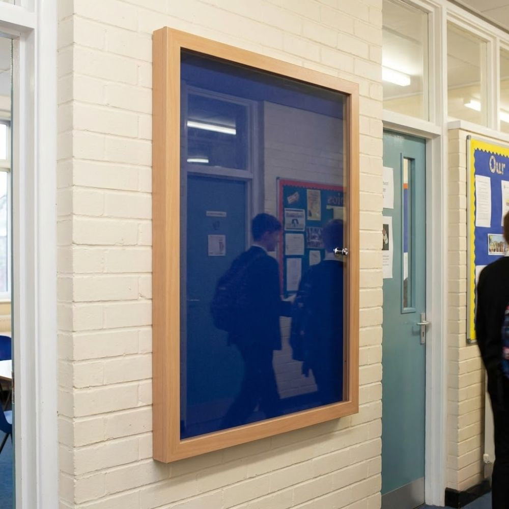 Eco-friendly 900 x 1200mm blue felt lockable noticeboard with wood effect frame and glazed door, wall mounted in a school corridor.