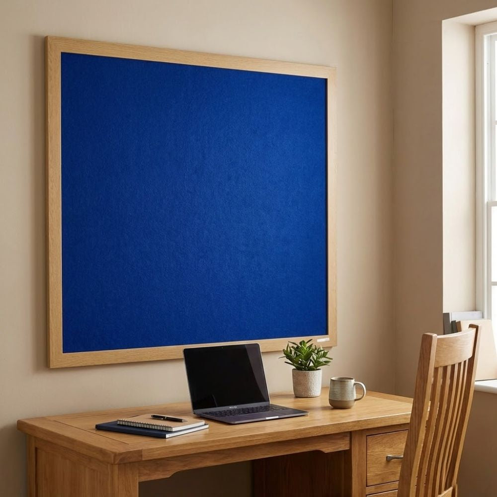 Eco-friendly 1200 x 1200mm blue felt noticeboard with wood effect frame, wall mounted above a wooden desk with laptop and chair in a bright home office.