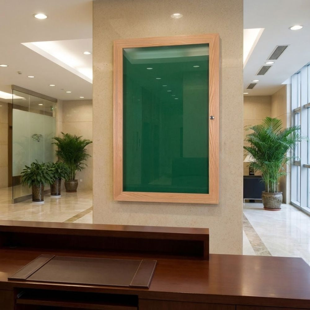 Eco-friendly green felt lockable noticeboard with wood effect frame and glazed front, displayed in a modern lobby area with tiled floor and indoor plants.