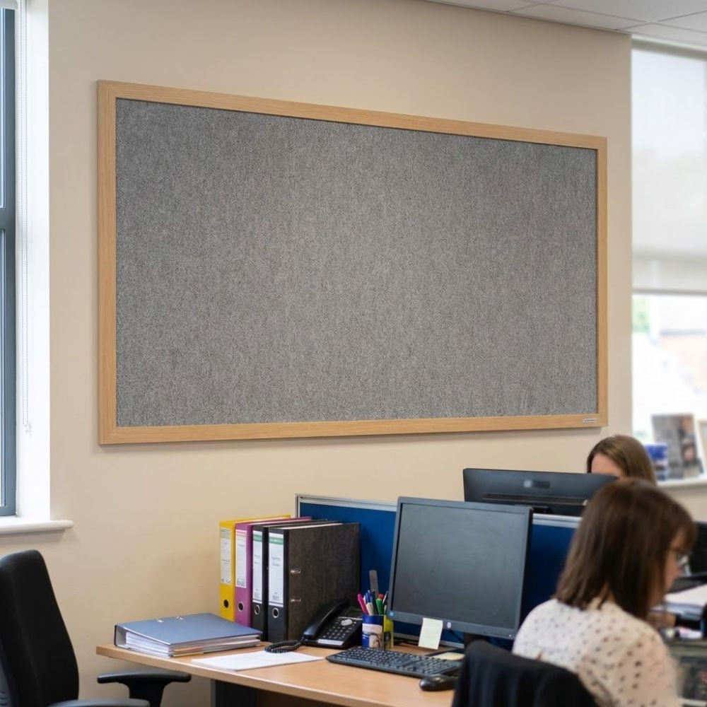 Eco-friendly 2400 x 1200mm grey felt noticeboard with wood effect frame, displayed in an open plan office above desks with computers and office staff.