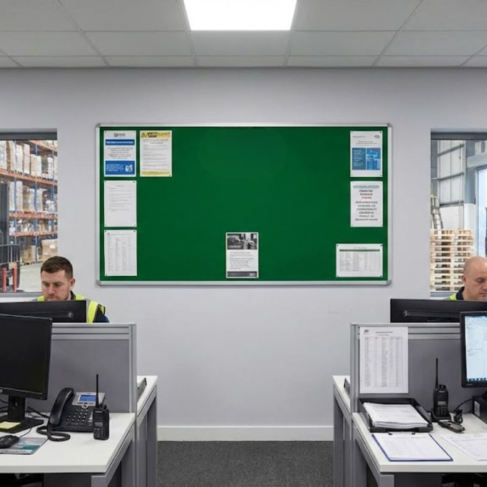 Extra large green aluminium framed noticeboard mounted on an office wall in a warehouse environment with staff desks and computer workstations positioned below.