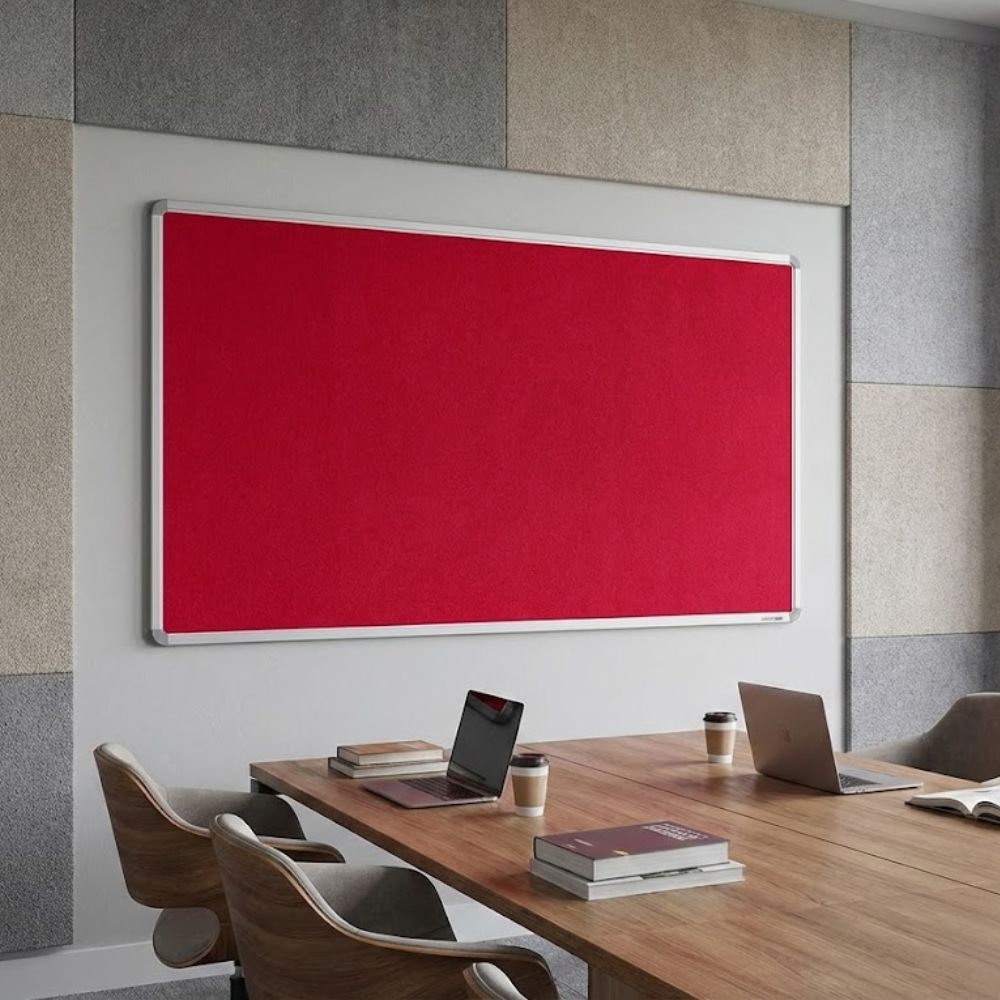 Extra large red aluminium framed noticeboard installed on an acoustic panel wall in a modern meeting room above a wooden conference table with laptops and chairs.