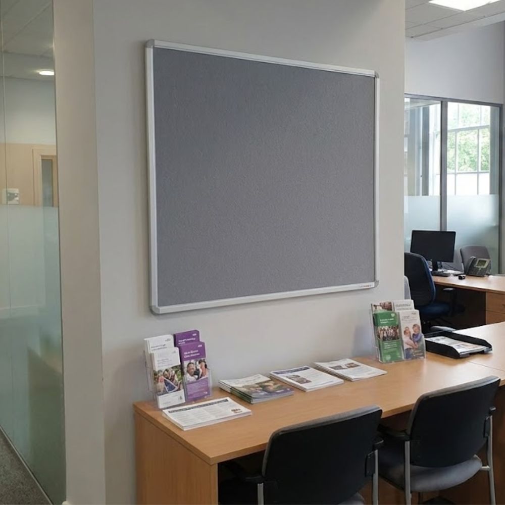 Grey aluminium framed noticeboard measuring 1500 x 1200mm installed on a wall in a reception area with desks, chairs and informational brochures displayed below.