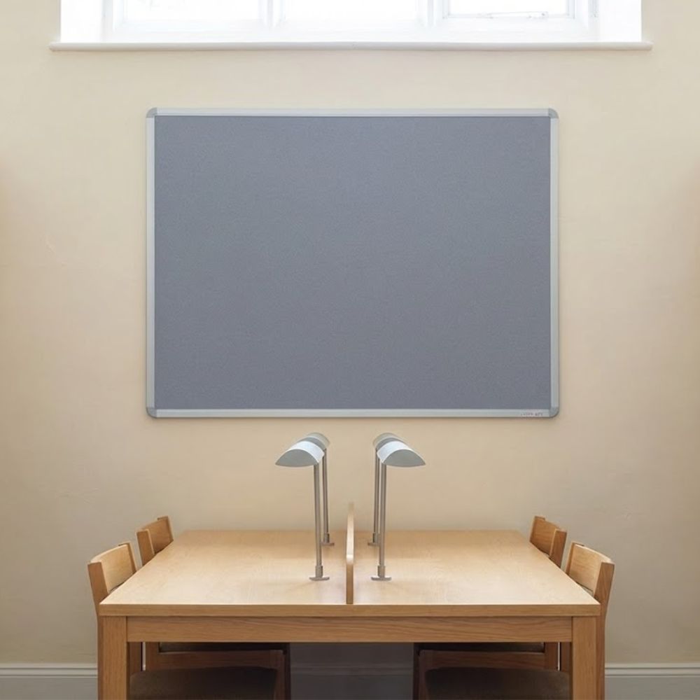Grey aluminium framed noticeboard measuring 1800 x 1200mm mounted on a wall above wooden study desks with lamps in a quiet study area.