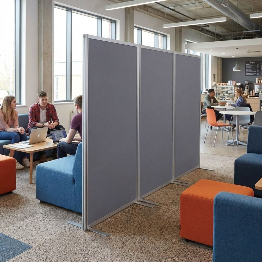 Grey panel aluminium freestanding display board used as a room divider in a modern breakout area with sofas, tables and people working on laptops.