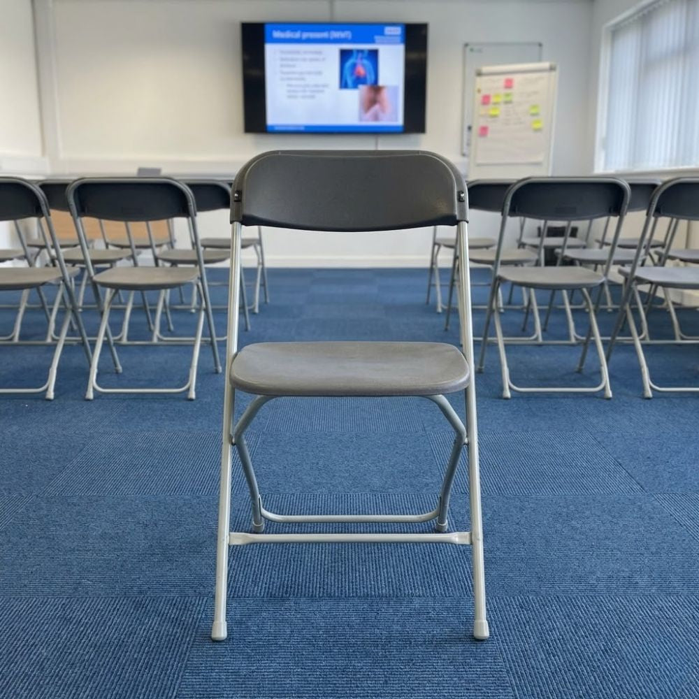 Grey folding assembly chair with metal frame, positioned in a corporate training room with presentation screen and arranged seating.