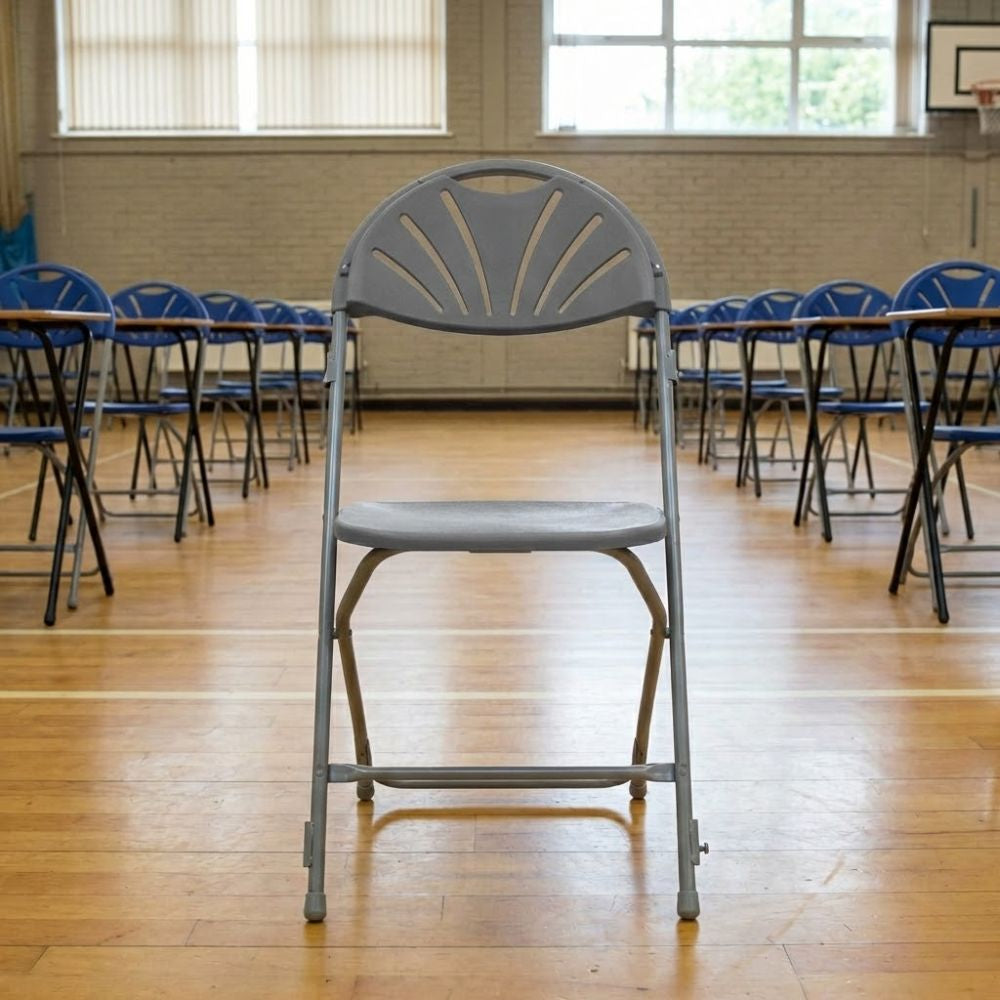 Grey plastic folding assembly chair with metal frame, placed at the front of rows of chairs and tables in a school exam hall.