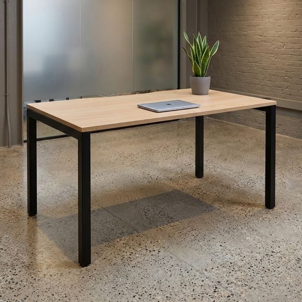 Simple wooden office desk with black metal legs, placed in a modern industrial office setting with concrete flooring, laptop, and potted plant.