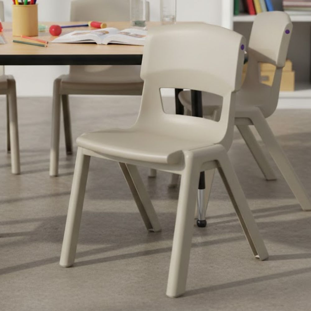 A sturdy light‑grey plastic school chair pulled up to a low activity desk in a school classroom environment, with art supplies and learning resources visible on the table.
