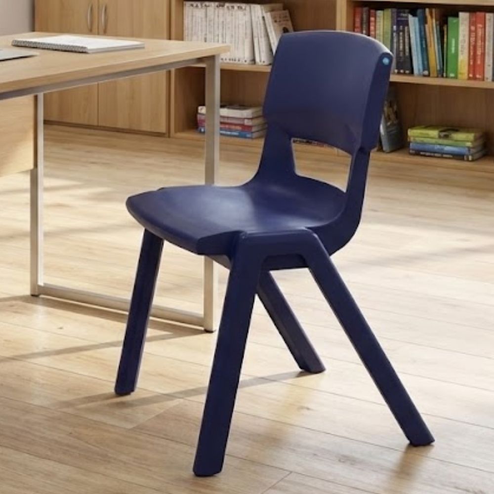 A navy blue plastic classroom chair arranged in a quiet study room with open bookshelves, a modern desk, and light wood flooring for a calm academic setting.