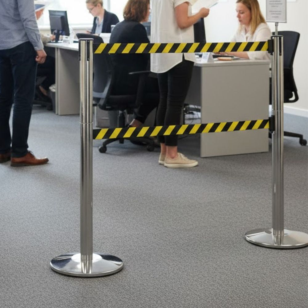 Polished steel retractable barrier post with twin 3.4m black and yellow chevron hazard belts for queue control in a busy office or reception environment.