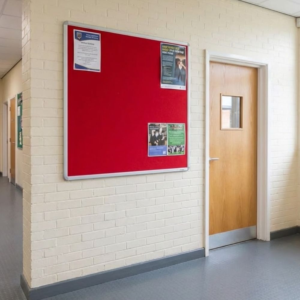 Red aluminium framed noticeboard 1200 x 1200mm mounted on a brick corridor wall with informational posters displayed beside a classroom door.