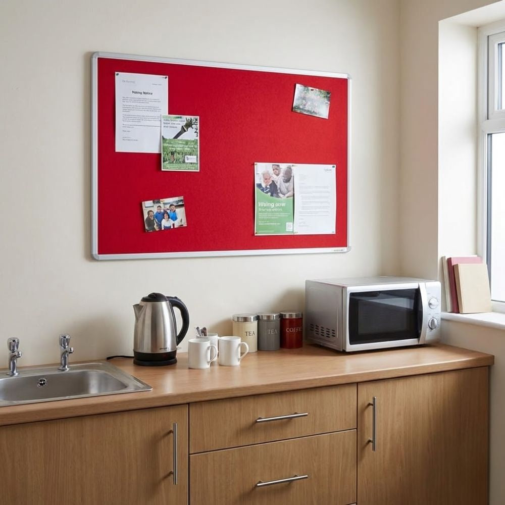 Red aluminium framed noticeboard mounted above a kitchenette counter with sink, kettle and microwave, displaying various workplace notices.