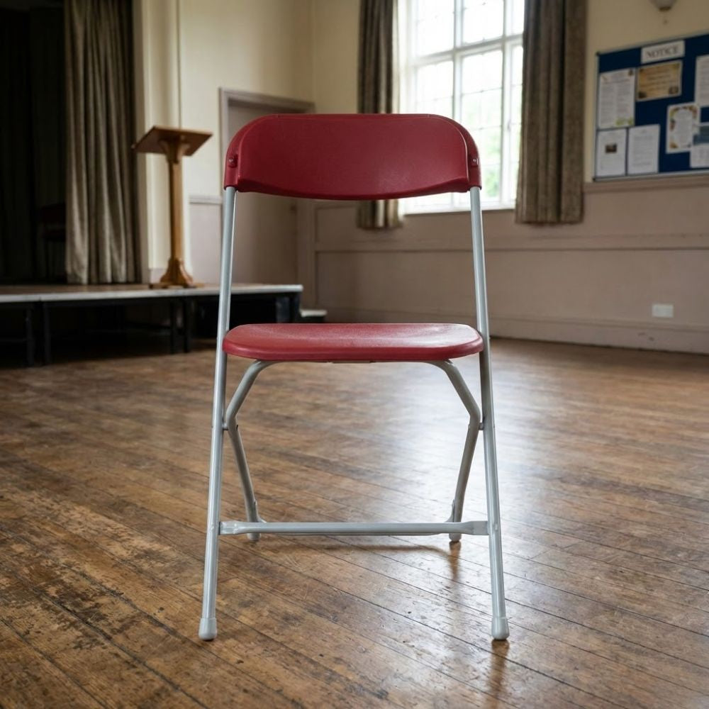 Red folding assembly chair with metal frame, placed on a wooden floor in a community hall with stage and noticeboard behind.
