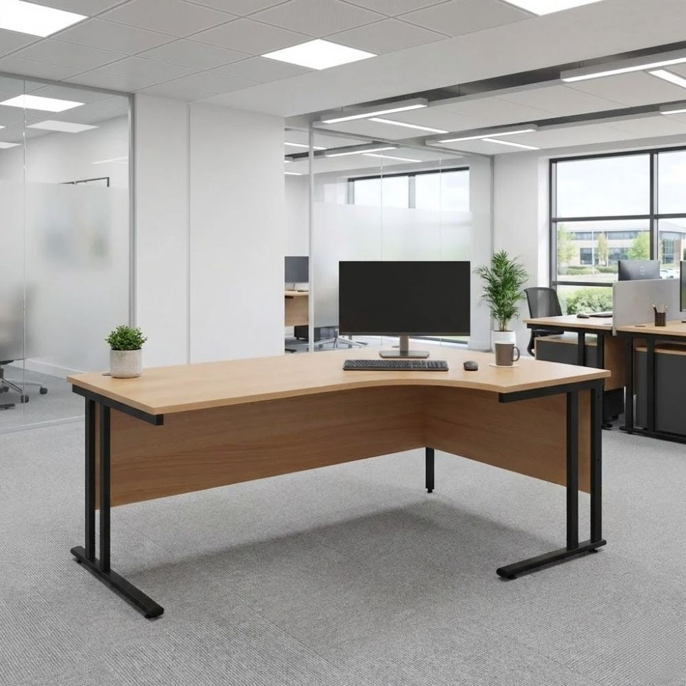 Right-hand return office desk with a spacious wood-finish worktop, black cantilever legs, and modesty panel, shown in a carpeted open-plan office with monitor and keyboard setup.