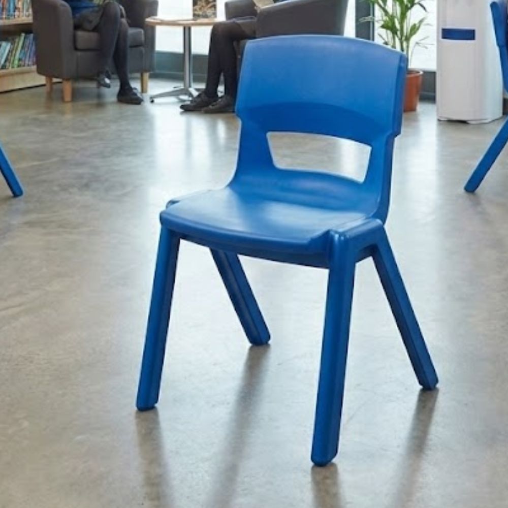 A royal blue plastic classroom chair in a sixth form or secondary school library lounge, set against a polished concrete floor with students seated in the background.