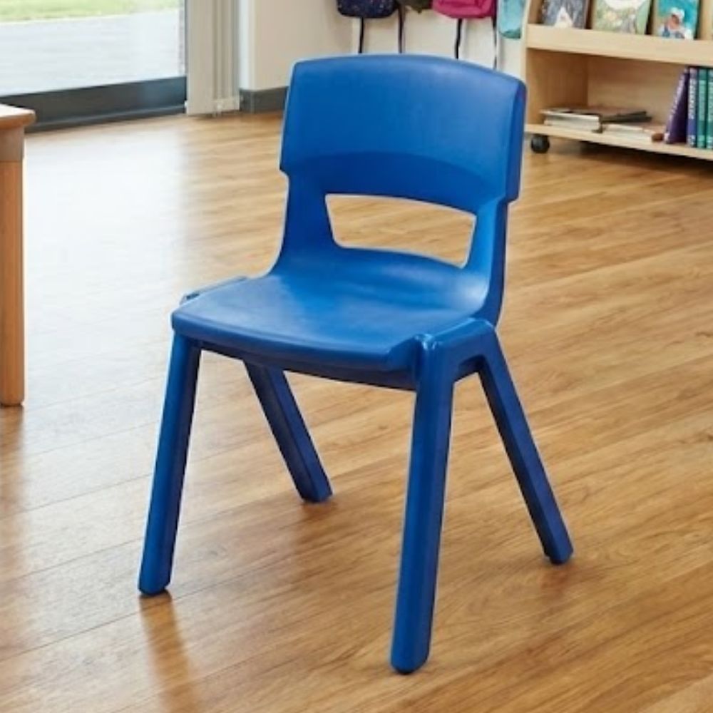 A durable royal blue primary school chair set in a sunlit classroom with natural wooden flooring and wall-mounted coat pegs in the background.