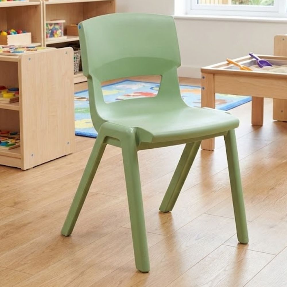 A sage green plastic chair for toddlers, placed in a light-filled nursery space surrounded by toy storage, a sand table, and soft rugs for interactive play.