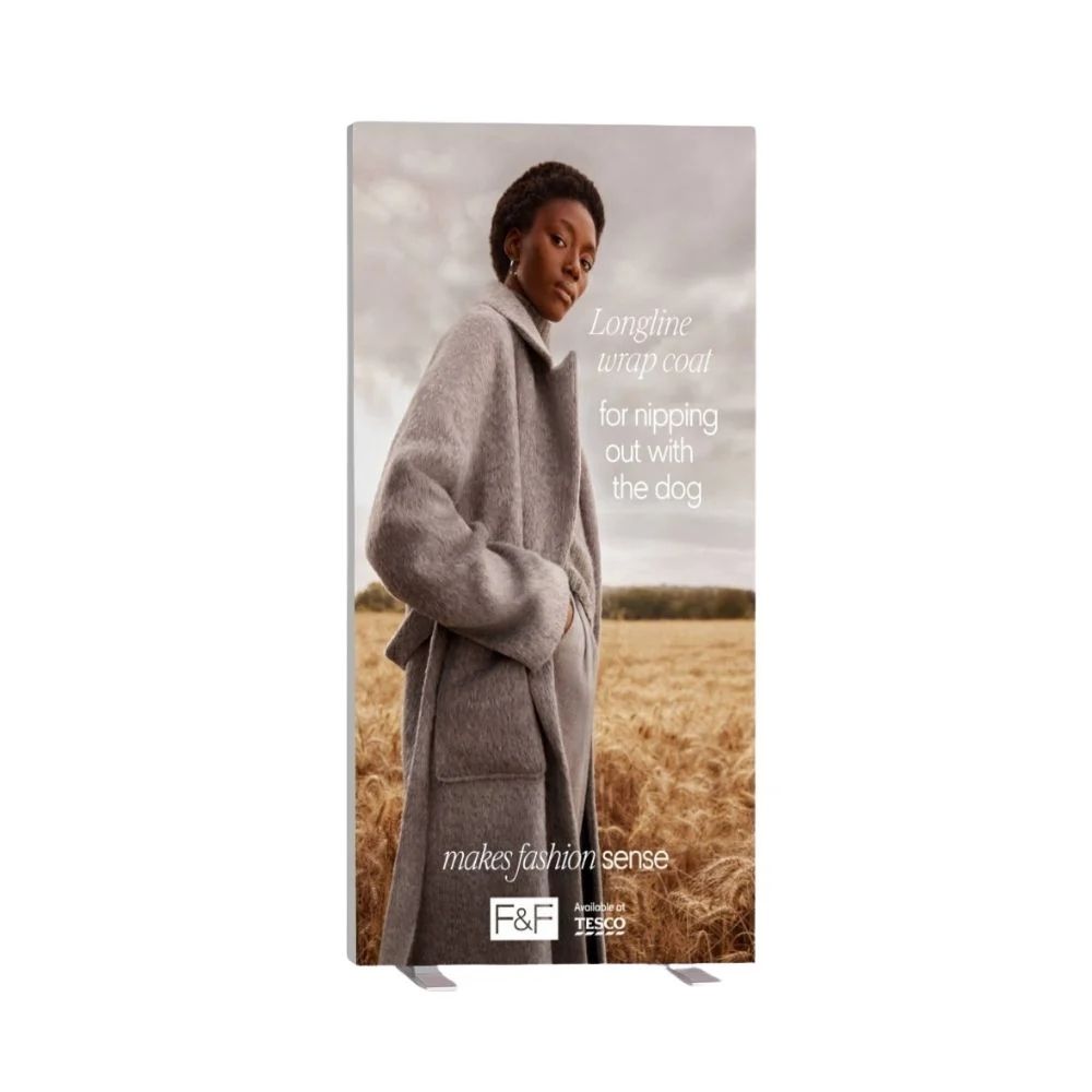 Tesco F&F fashion banner featuring longline wrap coat in grey, styled for outdoor dog walking in a golden field with cloudy sky backdrop.