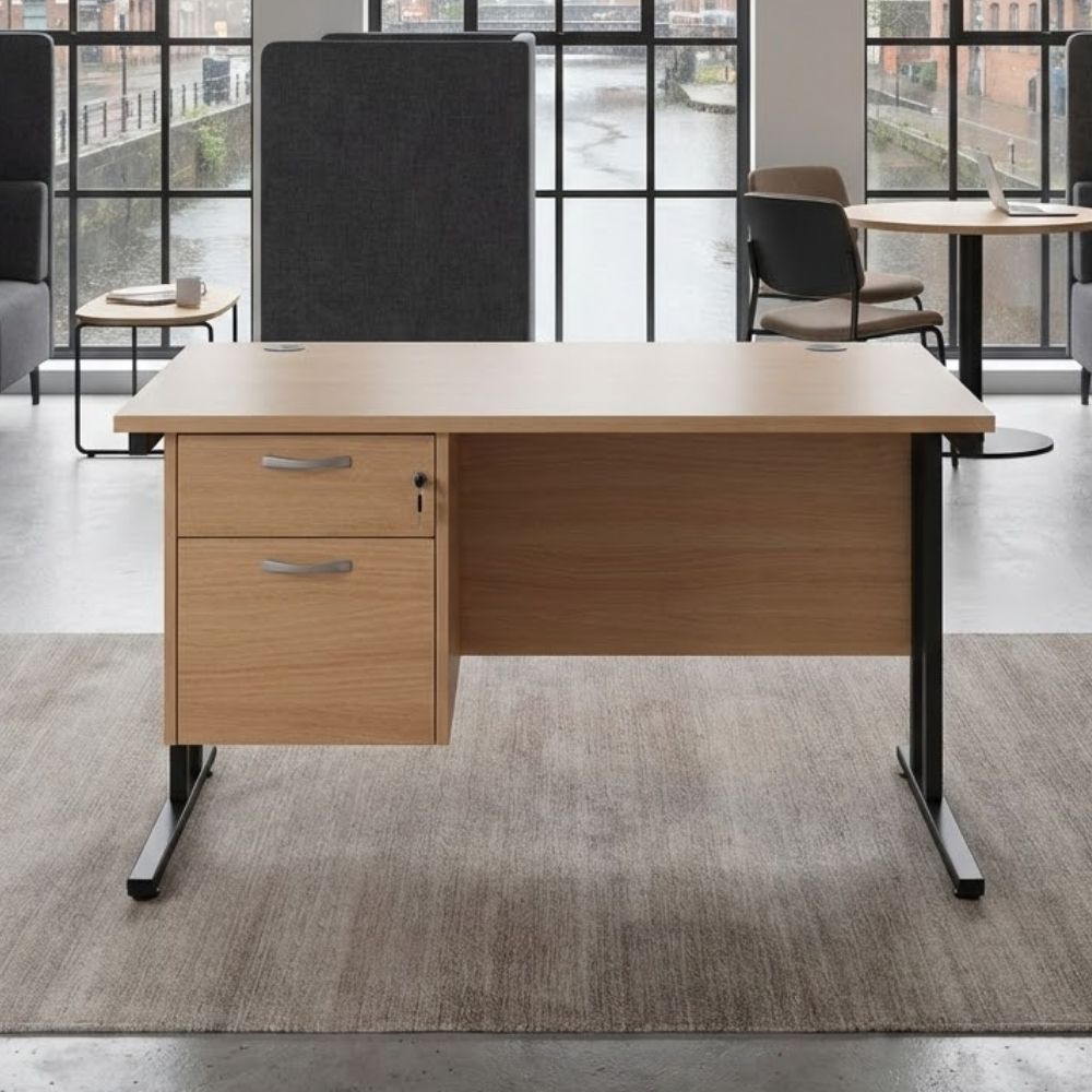 UK-made wooden office desk with modesty panel and two-drawer pedestal, black cantilever legs, shown in an industrial-style office with large factory windows and modern seating.