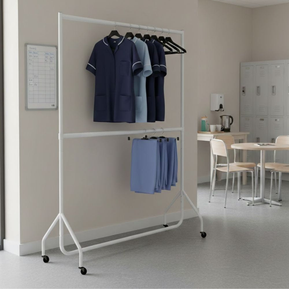 White 6ft two-tier mobile clothes rail holding nursing uniforms and trousers on hangers in a healthcare staff room with lockers and seating.