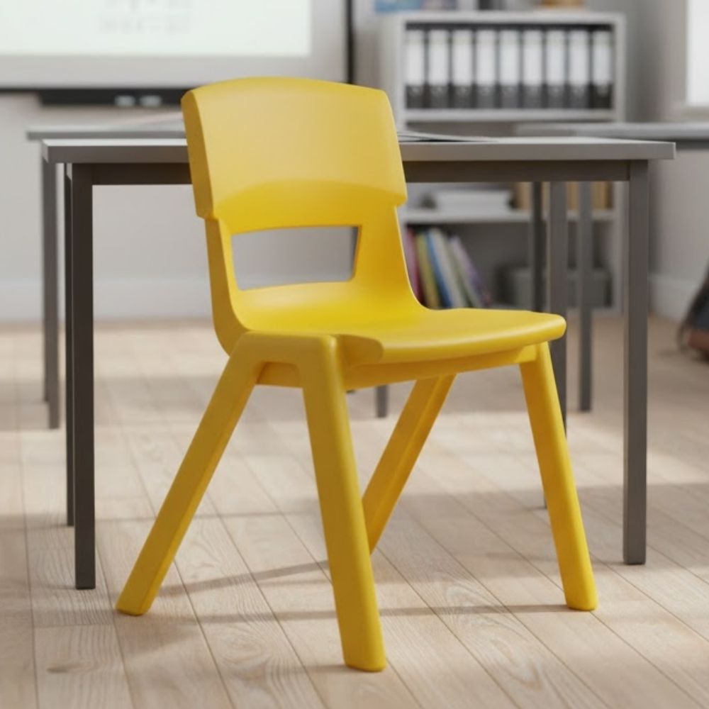 A yellow plastic chair with angled legs positioned in front of a desk in a modern classroom, with bookshelves and a projector screen behind.